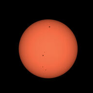 The orange disk of the sun with several dark sunspots